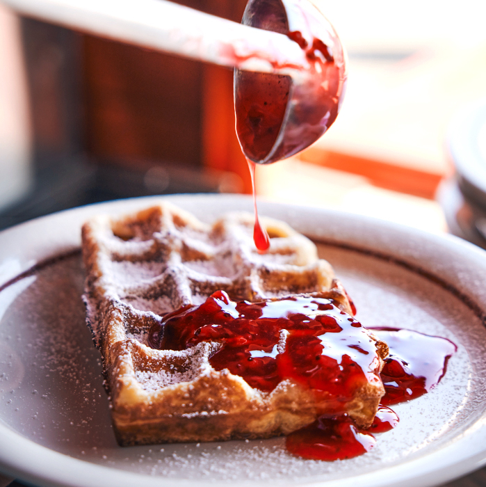 Frisch gebackene Waffel mit Puderzucker und warmer roter Fruchtsauce, die mit einer Kelle darüber gegeben wird – serviert in der Waffelhütte auf der Blockbräu Hafenterrasse.