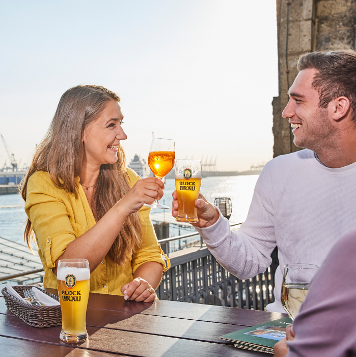 Zwei Gäste stoßen bei Sonnenschein mit Blockbräu Bier und einem Aperitif auf der Hafenterrasse an. Im Hintergrund sind die Elbe und der Hamburger Hafen zu sehen.
